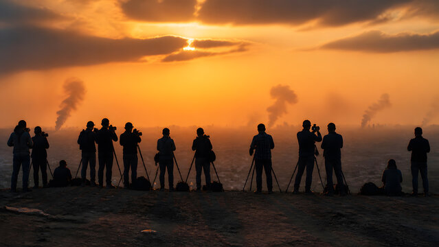Photographer group standing on hill with tripod and camera capturing smoke over city at orange sunset