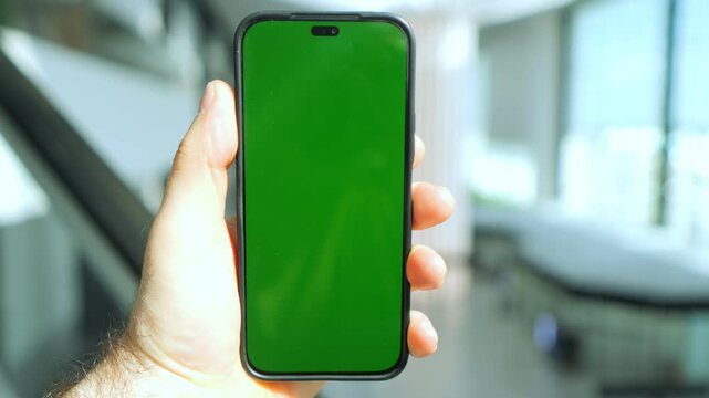 Hand holds smartphone with green screen. Background shows blurred indoor setting. Three keyframes show same scene from slightly different angles. Phone has black frame and no visible brand