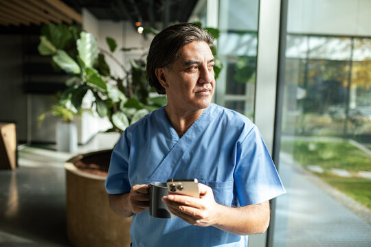 Healthcare worker in scrubs holding phone and coffee by window