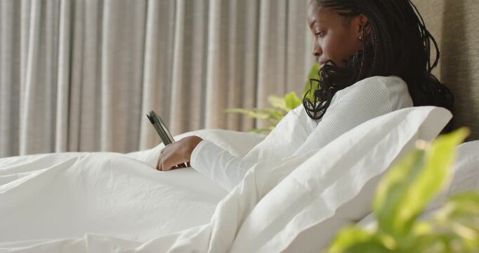 Adult African woman white top reclining in bed, scrolling then placing red phone on bedside table