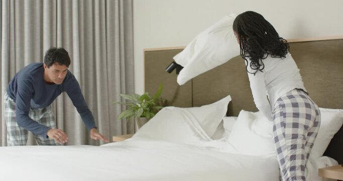 African American couple in pajamas preparing king bed in bedroom, fluffing pillows, smoothing duvet