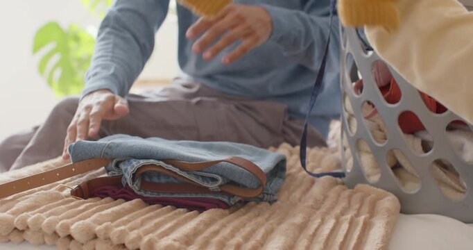 Hands are folding and stacking clothes on bed by window, starting with burgundy top for storage