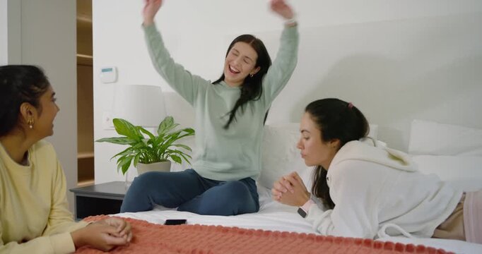 Diverse female friends sitting on bed, center woman seeing news on phone, dancing, causing claps