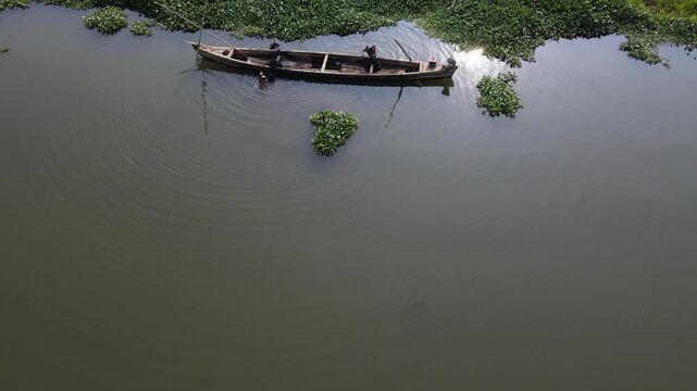 Aerial view of a wooden boat with traditional sand miners on the Serayu River near Kalirajut with water hyacinths and sunlight reflections in Patikraja, Central Java, Indonesia.