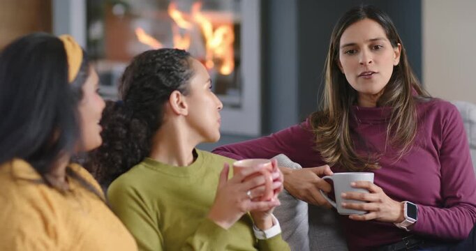 Diverse female friends sitting on sofa by lit fireplace, holding mugs, conversing and gesturing