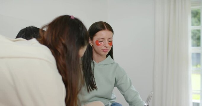 Female friends leaning at vanity urging seated woman with patches lifting orange jar, sniffing