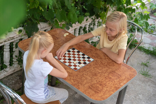 Young blonde boy and girl sitting at a table outdoors, engaging in a game of checkers, symbolizing childhood leisure and fun activities together during summer