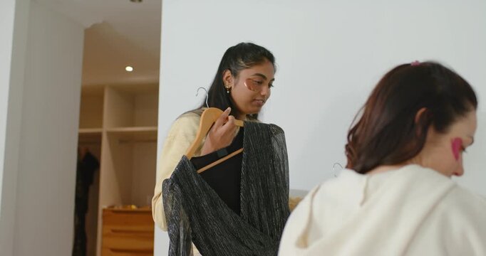 Diverse women Indian woman with hanger showing sequin dress to seated hoodie pal helping pick look