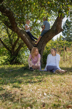 Four people spending time outdoors with two women relaxing on grass and two children sitting on tree branches, highlighting leisure and intergenerational bonding in nature