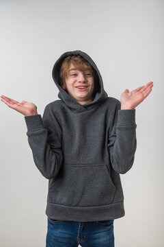 Young boy standing wearing a gray hoodie, shrugging shoulders with open hands and a cheerful expression, feeling uncertain against a clean studio background