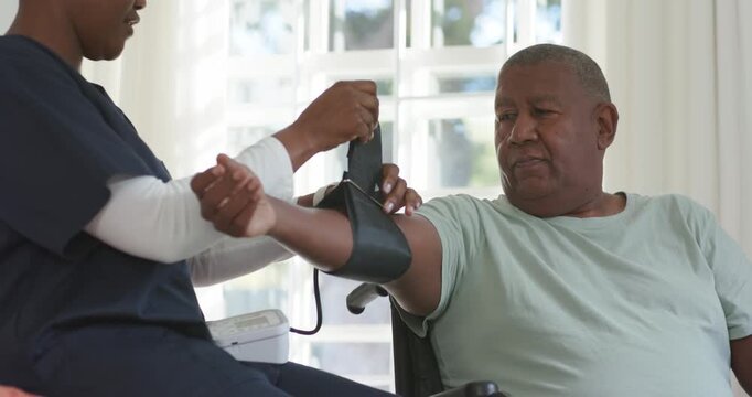 African American nurse fitting BP cuff, man extending arm in chair by window, checking BP monitor