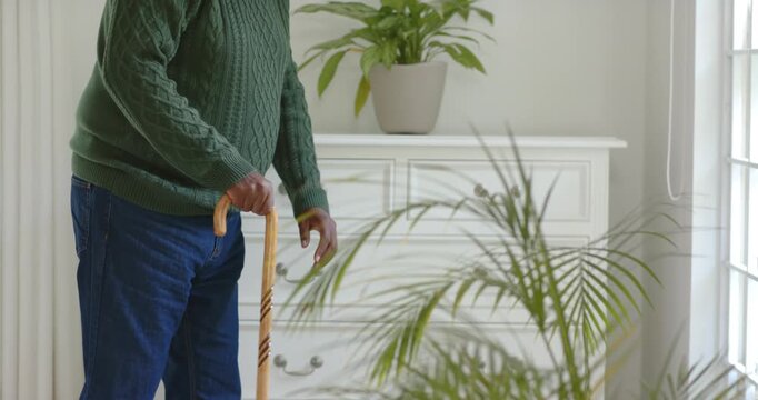 Senior African American man in green sweater at home by dresser, securing cane and peering outside