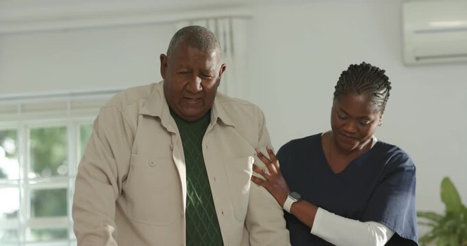 African American caregiver navy scrubs helping senior man stand, walk across room toward sofa