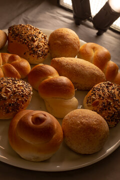 Assorted freshly baked bread rolls and seeded buns sit on white plate, showing golden crust texture under warm inviting light for meal and snack ideas.