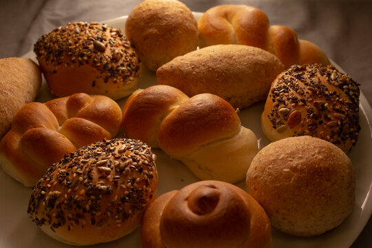 Close view of mixed bread rolls on white serving plate showing seeded rolls, plain rolls, and twisted rolls under warm lighting with visible crust and soft crumb texture.