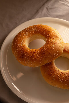 Top-down close-up of sesame-topped bagel on white ceramic plate, showing golden crust texture, visible crumbs, soft directional light, and blurred fabric background.
