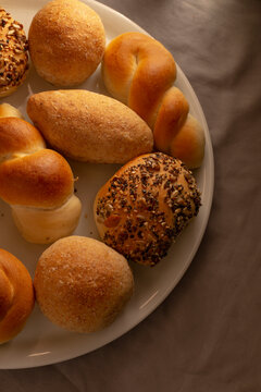 Close-up overhead view of assorted baked bread rolls on white plate showing seeded and braided shapes, plus plain rolls, on neutral cloth background.