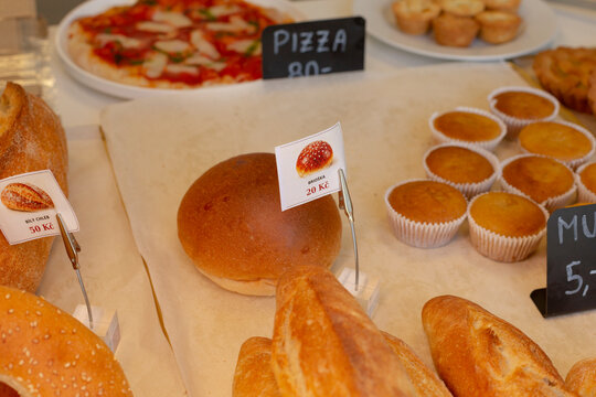 Close-up of assorted breads and muffins on bakery counter, showing brioche rolls, seeded loaves, and paper-cup treats beside small printed price tags.