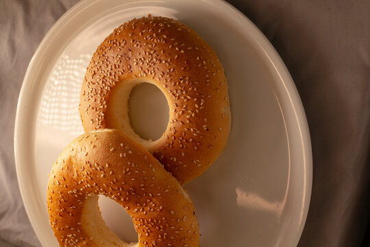 Two sesame seed bagels sit on white ceramic plate in tight top view, showing golden crust texture and scattered seeds against neutral cloth background.