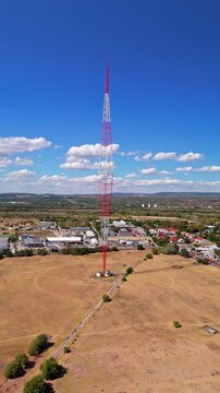 Drone view of the Lakihegy radio mast rising above dry fields and suburban development in Szigetszentmikl&oacute;s, Hungary, under a blue sky, with guy wires stretching across open terrain.