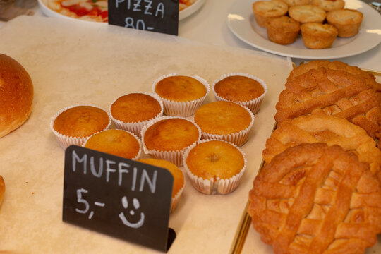 Close view of plain muffins baked in white paper cups arranged on bakery display counter, with adjacent baked pies and chalkboard price sign partially visible.
