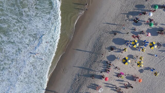 Top-down aerial view of colorful umbrellas and beachgoers on Copacabana Beach as waves roll onto the sand, Rio de Janeiro, Brazil.