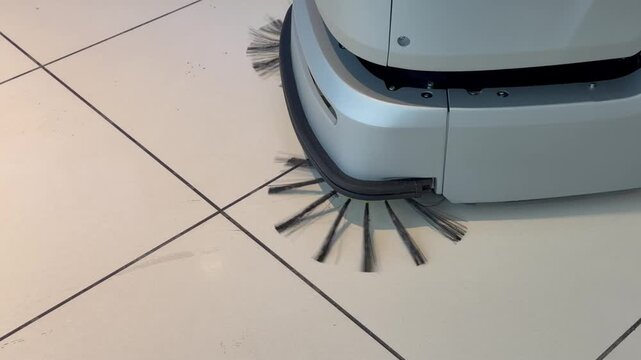 Modern autonomous cleaning robot with rotating brushes sweeping white tiled floor indoors. Smart automated floor scrubber machine, janitorial service and commercial cleaning technology concept.