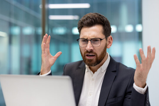 A close-up shot of a young, disappointed businessman sitting at a desk in an office, looking dejectedly at his laptop screen and spreading his hands