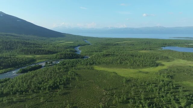 Aerial view of the Abisko National Park with the Kungsleden hiking trail in Sweden. A winding river flows through lush forests towards a vast lake, framed by snow-capped mountains.