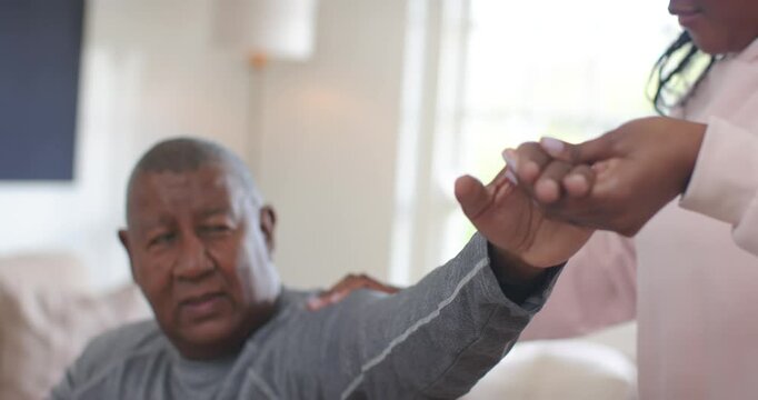 African American woman holding and turning senior man's hand on sofa, rubbing shoulder to ease pain