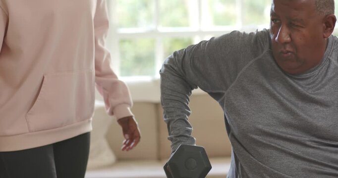 Senior man lifting hex dumbbell at home, woman in hoodie approaching, steadying