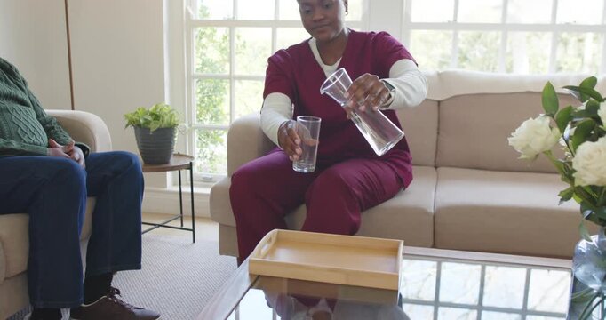 African American nurse pouring water from carafe into glass for senior after request in living room
