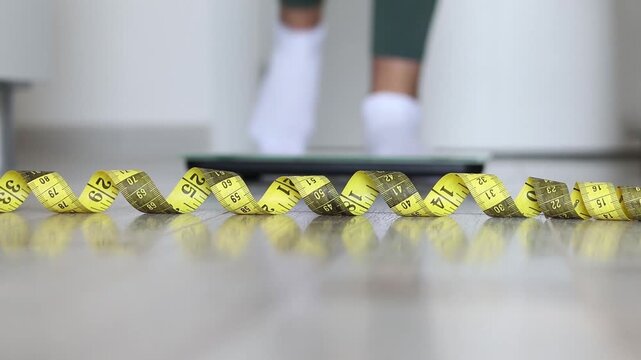 Measuring tape on a floor with woman's feet standing on a weight scale in the background. Wellness, fitness, and slimming concept.