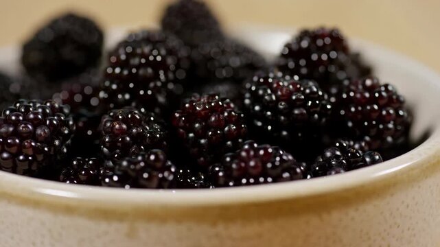 Blackberries piled in white bowl with glossy surfaces in close-up food scene, showing detailed berry clusters, dark colors, soft light, and fresh ingredient presentation with shallow depth composition