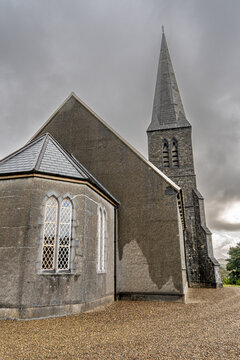Vertical shot of a gothic revival stone church featuring a tall spire, leaded glass windows, and stone apse on gravel ground under a dramatic cloudy sky in Ireland.
