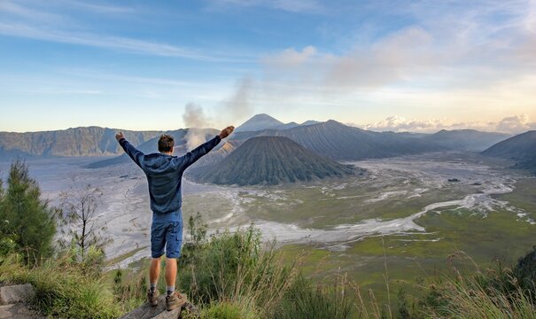 Young man stretches arms in the air, volcanic landscape, view in Tengger Caldera, smoking volcano Gunung Bromo, in front Mt. Batok, in the back Mt. Kursi, Mt. Gunung Semeru, National Park Bromo-Tengger-Semeru, Java, Indonesia