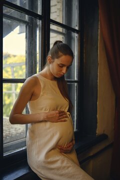A pregnant woman stands by a window in her home, cradling her baby bump and gently talks with the baby in the womb