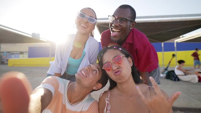 Group of four young, multi ethnic friends laughing and making funny faces while taking a selfie together outdoors on a sunny day, showcasing a moment of genuine joy and youthful energy