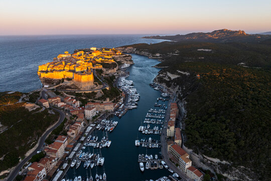 Idylllic drone view of Bonifacio old town illuminated at sunrise with boats moored at the dock of the fjord-shaped harbour, south of Corsica (Corse-Du-Sud), Corsica, Mediterranean Sea, France