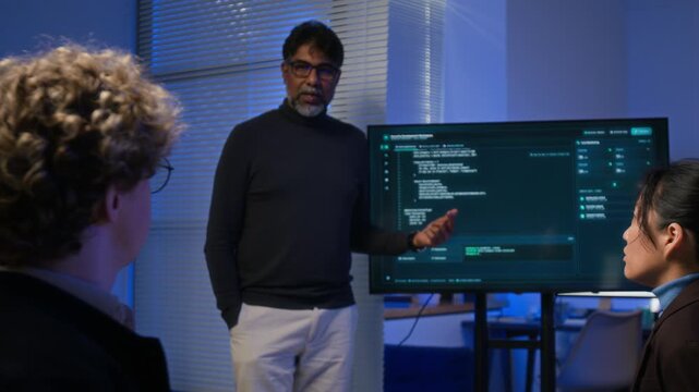 Middle aged South Asian programmer explaining code on screen to diverse colleagues in tech office. Footage ideal for corporate training videos, IT tutorials and cybersecurity advertising