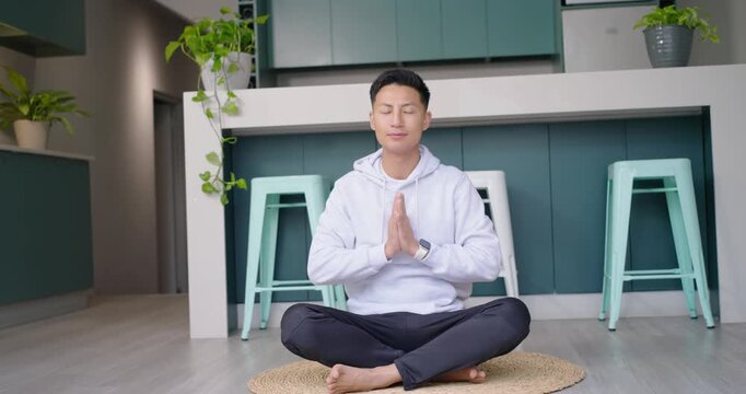 Mid-adult Asian man sitting on mat in kitchen, starting meditation with hands pressed, breathing