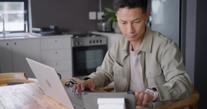 Asian man in 30s starting work session at kitchen table, using laptop and tablet