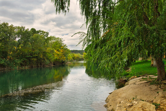 View of the Tagus River in Zorita de los Canes, Guadalajara, Castilla-La Mancha, Spain. Overhanging willow branches frame the water, with reflections of riverside trees, a rocky bank and a cloudy sky.