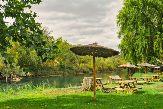 Wooden picnic tables and thatched parasols beside the Tagus River, surrounded by willow trees in Zorita de los Canes, Guadalajara, Castilla-La Mancha, Spain.