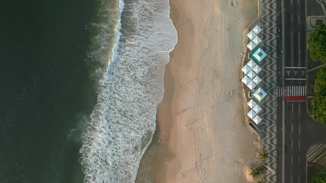 Aerial view of the iconic wave-patterned sidewalk, white tents, and ocean waves along the sandy beach in Rio de Janeiro, Brazil.
