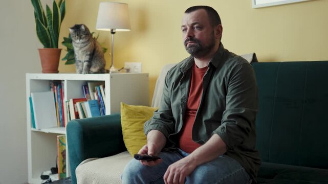 Middle-aged man sitting straight on sofa pointing remote control at screen