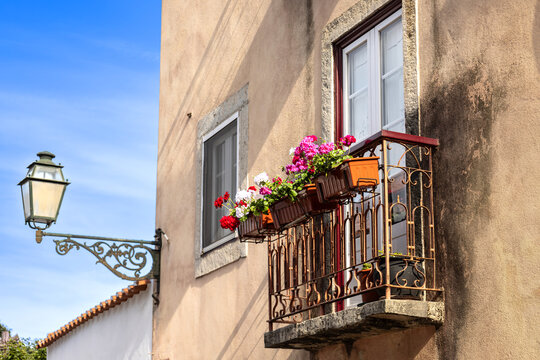 View of Alfama traditional architecture featuring a wrought iron balcony with vibrant pink and white geraniums and an ornate street lamp on a building in Lisbon, Lisbon, Portugal.