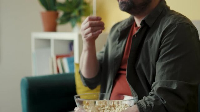 Mid-section of relaxed adult male holding bowl of popcorn while sitting on couch
