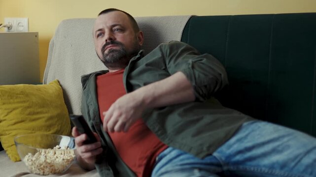 Reclining man pointing TV remote control while resting on sofa with popcorn