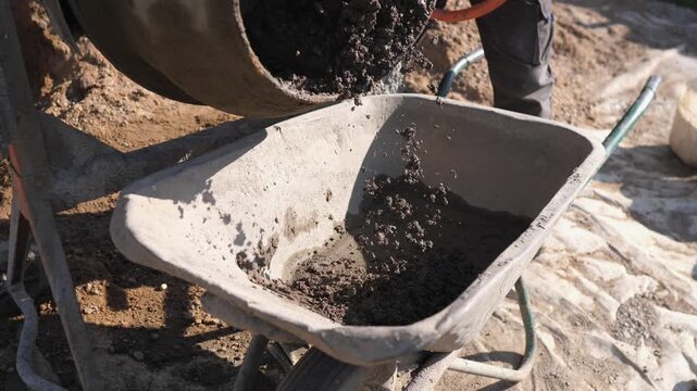 Wet concrete falls from rotating drum into wheelbarrow at dusty construction site, while worker stands nearby and sunlight highlights rough texture, active groundwork, and manual building work.